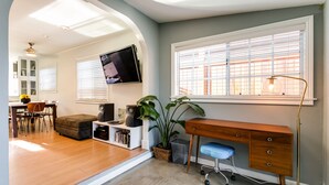 Una habitación con un escritorio de madera, una silla azul, un televisor montado en la pared y un ventanal con persianas.
