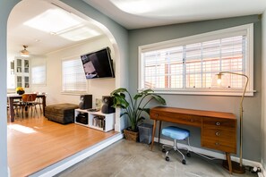 A room with a wooden desk, a blue chair, a television mounted on the wall, and a large window with blinds.