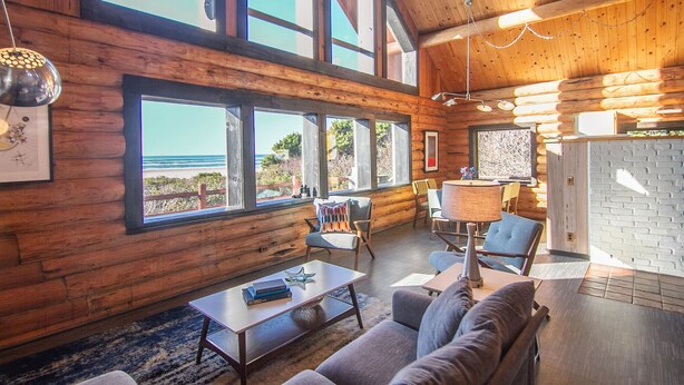 Amplia habitación con grandes ventanales con vista a la playa, paredes de madera, un rincón de descanso acogedor y una cocina moderna visible a través de un vano abierto.