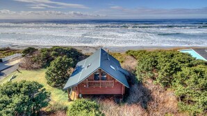 Una cabaña de madera con techo inclinado, rodeada de abundante vegetación y con vistas a una playa y sus olas.