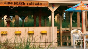 A beach bar with a wooden sign, outdoor seating, and a tropical umbrella.