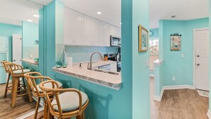 Une cuisine aux murs de couleur turquoise, avec des meubles blancs et un plan de travail en marbre. On y trouve trois tabourets de bar en osier.