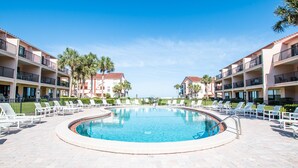 Une piscine circulaire entourée de chaises longues et de palmiers, elle-même bordée d’immeubles à plusieurs étages dotés de balcons.