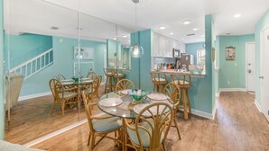 Un coin repas avec des tables rondes en verre et des chaises en osier, une cuisine avec des meubles blancs et un escalier avec des rampes blanches.