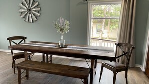 A dining area with a wooden table, two chairs, and a bench. A decorative metal piece is on the wall, and a window with curtains is visible.