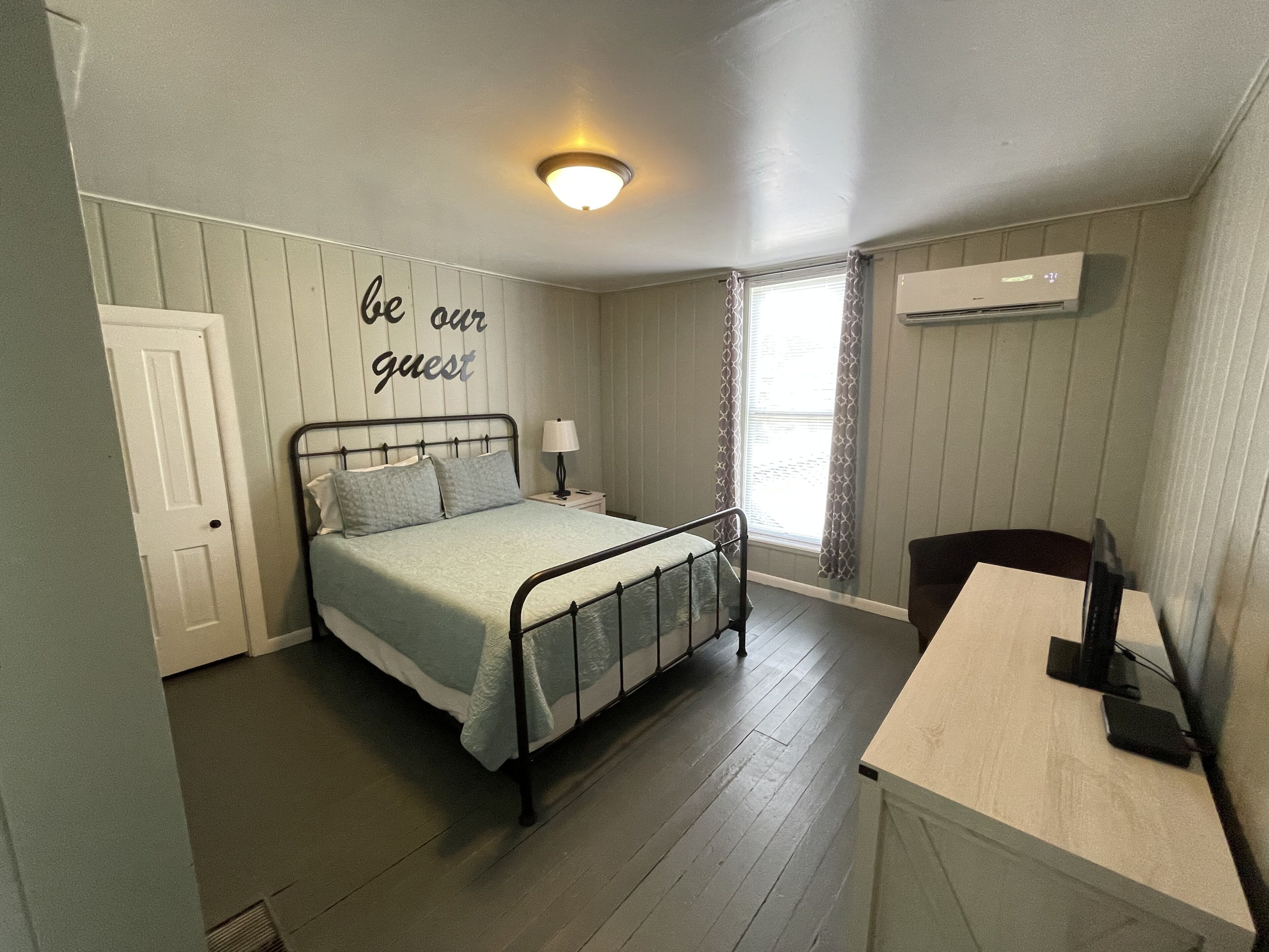 A bedroom with a bed, a bedside table with a lamp, a desk with a chair, a window with curtains, and a wall with a sign saying "be our guest".