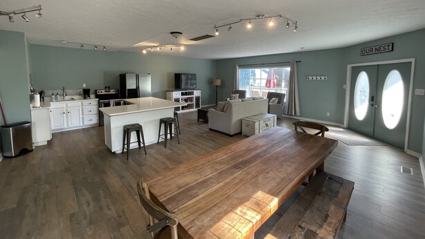 A spacious living area with a wooden dining table, a kitchen with white cabinets, and a large window providing natural light.