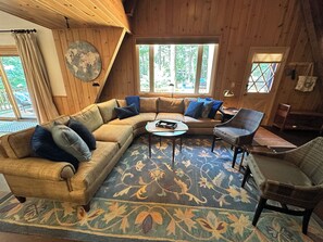 Ein gemütliches Zimmer mit einer großen Eckcouch, einem kleinen runden Tisch und einem Sessel. Große Fenster sorgen für natürliches Licht und Ausblick ins Freie.