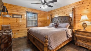 A wooden-paneled bedroom with a bed, a nightstand, a lamp, a ceiling fan, and a window.