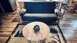 A room with a wooden floor, a blue sofa with patterned cushions, a round coffee table with a remote control, and a rug with a moose design. - Bear Den Log Cabin (Wetmore)