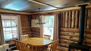 A wooden cabin interior with a round wooden table, four chairs, a chandelier, a map on the wall, and a wood-burning stove. - Bear Den Log Cabin (Wetmore)