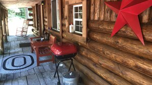 A wooden cabin with a porch, a rocking chair, a table, a red grill, and a metal bucket. - Bear Den Log Cabin (Wetmore)