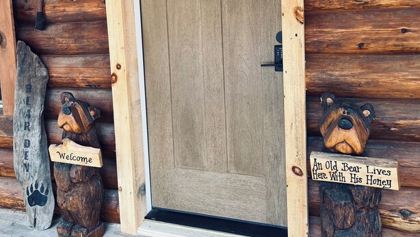 A wooden cabin door with a welcome sign and two bear sculptures, one with a sign reading "Welcome" and the other "An Old Bear Lives Here With His Honey." - Bear Den Log Cabin (Wetmore)