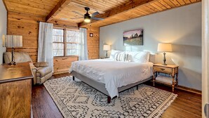 A bedroom with a wooden ceiling, a bed with white bedding, a desk with a lamp, and a patterned rug.