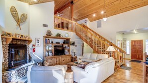 A spacious living room with a fireplace, white leather sofas, a wooden staircase, and a bookshelf.