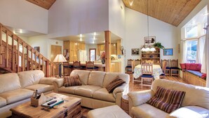 A spacious living area with a high ceiling, wooden beams, and a staircase. There are multiple seating areas with leather sofas and a dining table.