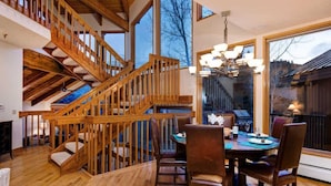 Una escalera de madera que lleva a un nivel superior, un área de comedor con una mesa y sillas, y ventanas grandes con vistas al exterior.