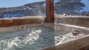 Un jacuzzi con un poste de madera en el centro, rodeado de árboles cubiertos de nieve y montañas al fondo.