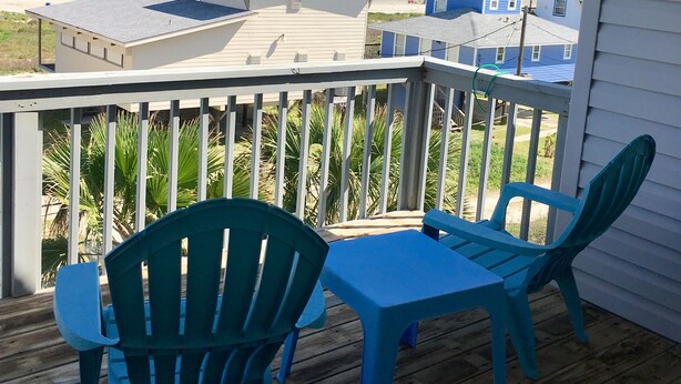 Una terraza de madera con dos sillas azules y una mesa, con vista a una playa y casas al fondo.