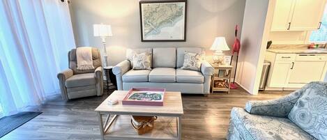 A living room with a grey sofa, a patterned armchair, a coffee table, and a map on the wall.