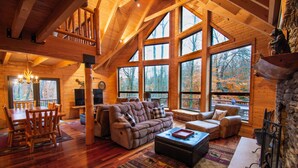 A spacious living room with a large sofa, a coffee table, and a fireplace. There are large windows offering a view of the forest. - Stream Front Log Home near 7 Springs, 4 BR (Rockwood)