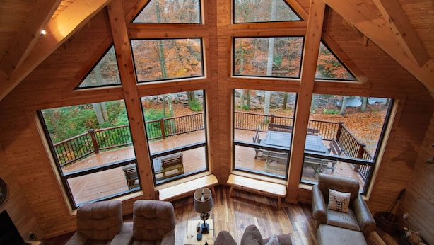 A high-ceilinged room with large windows, wooden flooring, and a view of an outdoor deck. - Stream Front Log Home near 7 Springs, 4 BR (Rockwood)