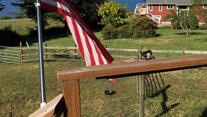 Una bandera estadounidense en un asta, una valla de madera, y una casa con techo rojo y molduras blancas, con vistas a un área cubierta de césped y al océano.