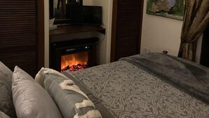 Un dormitorio con chimenea, una cama con ropa de cama estampada, un cuadro en la pared y contraventanas de madera.