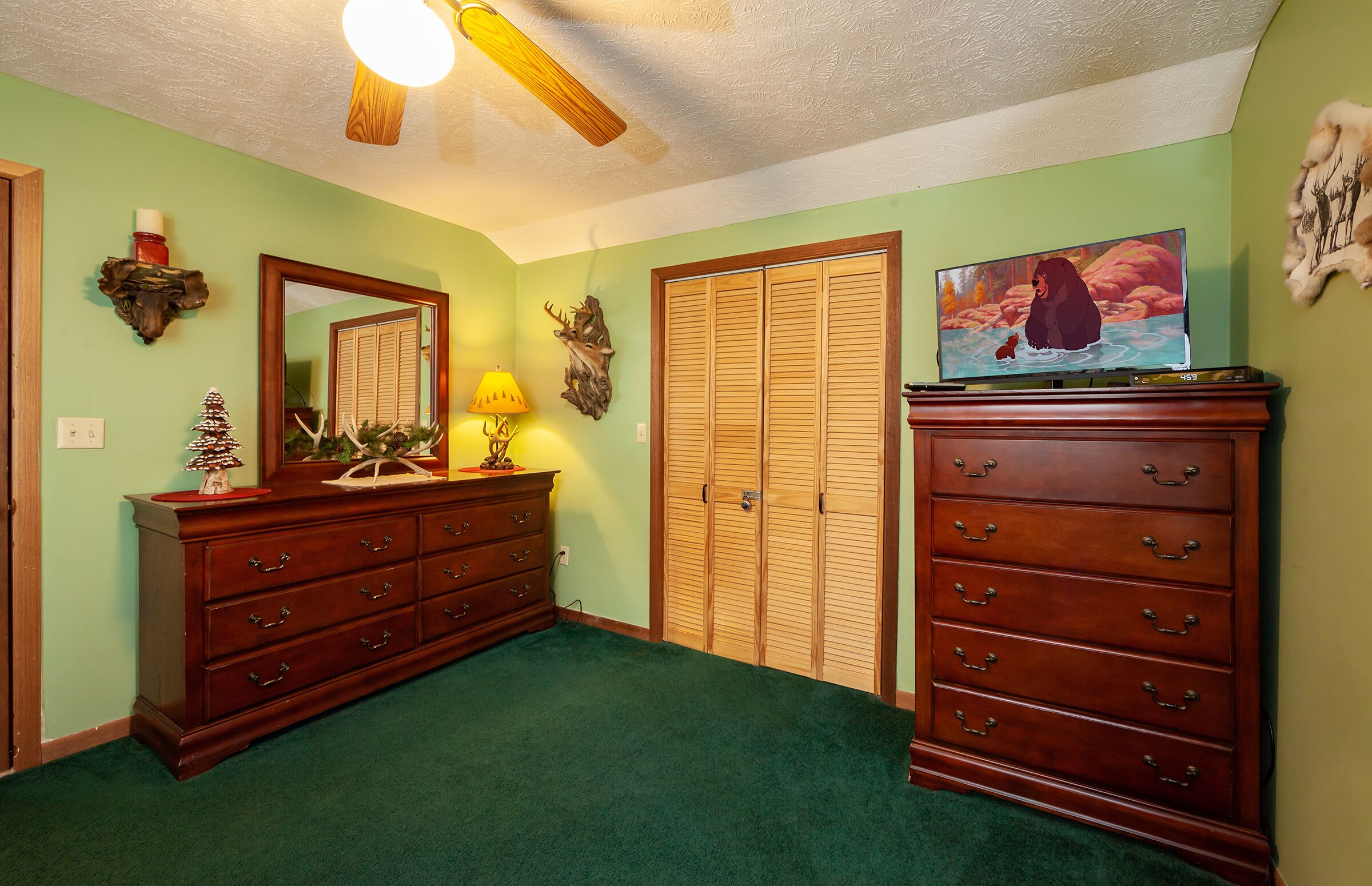 Een slaapkamer met een groen tapijt, een houten dressoir, een spiegel, een televisie en een plafondventilator.