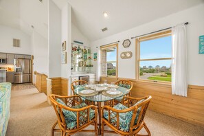 A dining area with a round glass table and wicker chairs, a kitchen with modern appliances, and a large window offering a view of the surrounding area.