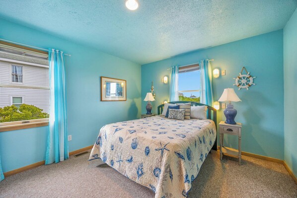 A bedroom with a bed, bedside tables, a window with curtains, a wall-mounted picture, and a ship's wheel decoration.