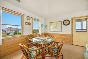 A dining room with a glass table surrounded by wicker chairs, a clock, and a framed map on the wall. - Soundfront Home w/ Hot Tub, Windsports Access, Community Tennis and Pickleball (Rodanthe)