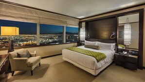 Ein Hotelzimmer mit einem großen Bett, einem Sessel, einem Nachttisch, einer Lampe und Blick auf eine nächtliche Skyline.