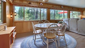 Habitación con paneles de madera, una mesa redonda y sillas, un sofá y ventanas amplias con vistas a árboles.