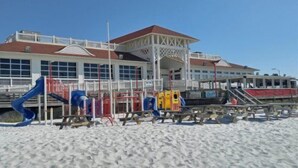 Una platja amb un parc infantil, un edifici amb teulada vermella i una torre de socorristes.