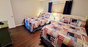 A room with two beds, each covered with a quilt featuring a patriotic pattern. There is a window with blinds and a lamp on a bedside table.