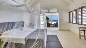 A bedroom with a bed under a mosquito net, a ceiling fan, and a view of the sea through large windows. - Oceanfront with private beach - 4 bedrooms, 4 bathrooms (Kralendijk)