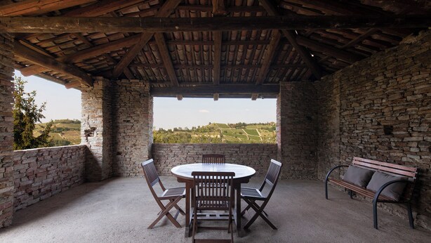 A covered outdoor patio with a stone wall, a round table with chairs, and a bench. - Breathtaking views, excellent food and wine: Barbaresco, Barolo, Alba truffles.. (Trezzo Tinella)