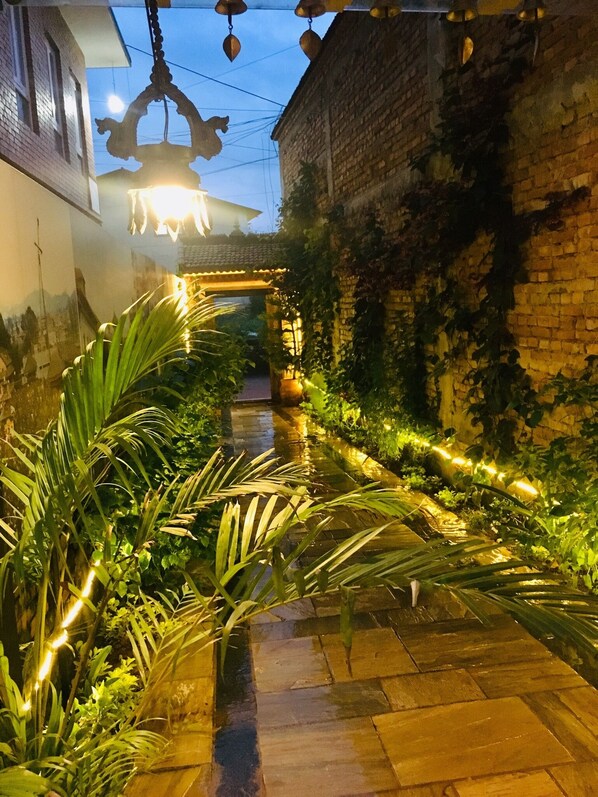 A narrow, illuminated pathway with a brick wall on one side and a building on the other, featuring a hanging light fixture and a small archway. - Hotel Central Bhaktapur (Bhaktapur)