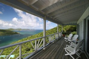 Eine Holzterrasse mit weißen Schaukelstühlen und Blick auf die Küste, mit Booten und Grünpflanzen.