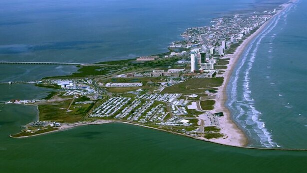 Vista aérea de uma cidade costeira com extensa faixa de areia, área urbana densa e um porto de iates.