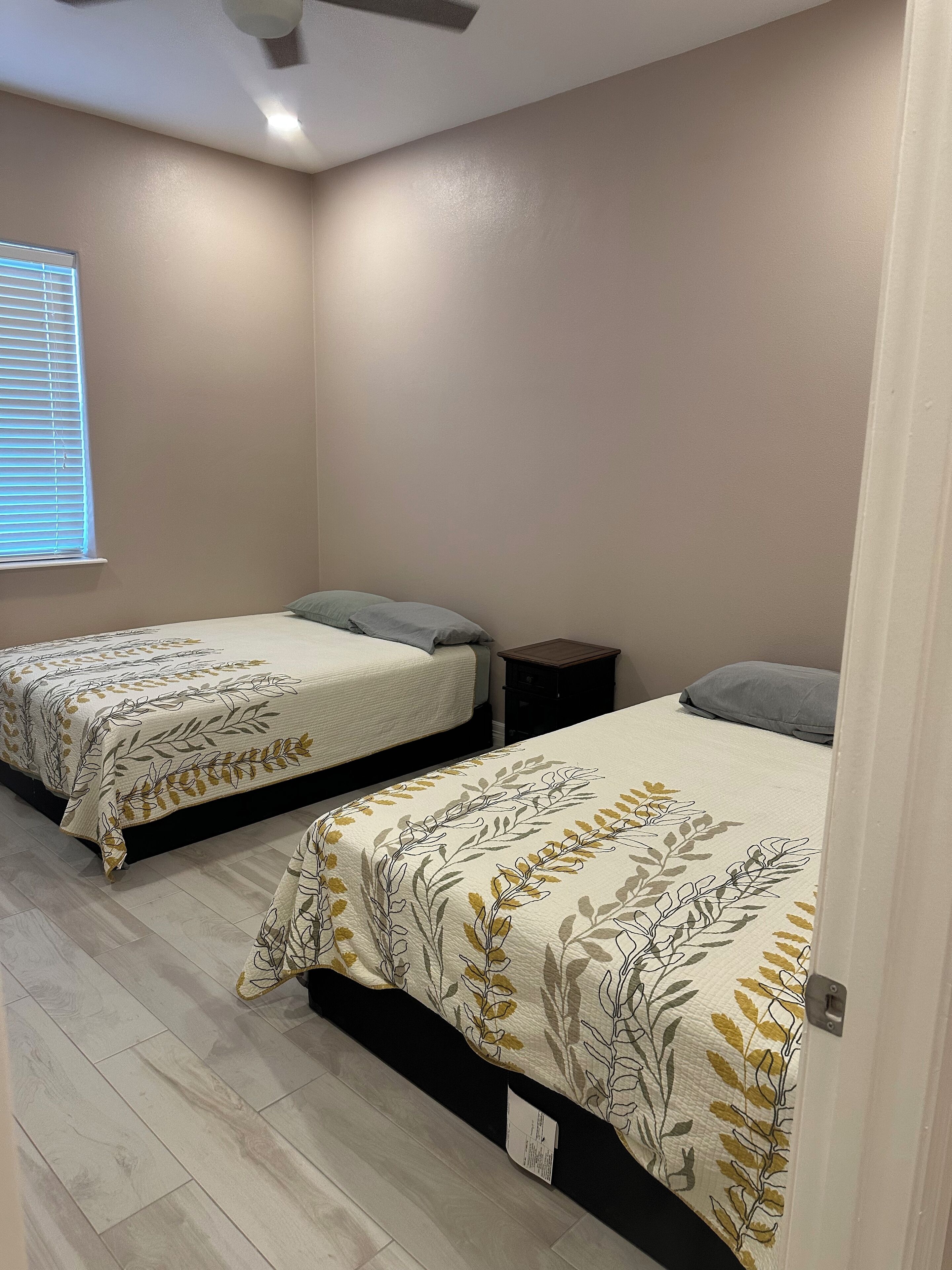 A bedroom with two beds, each with a floral bedspread, a small bedside table, and a window with blinds.