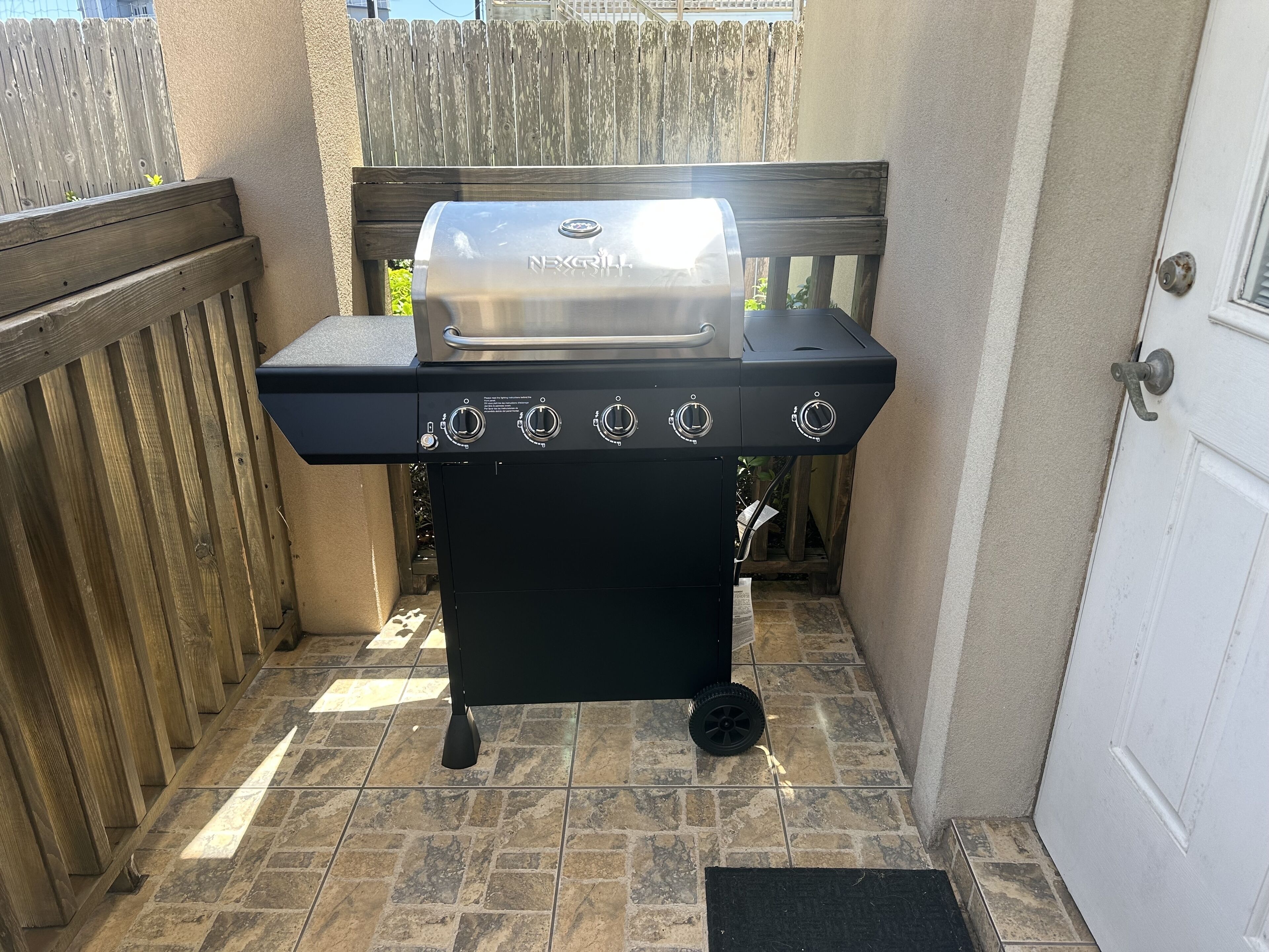 Una parrilla negra para exteriores con cuatro quemadores y pantalla digital, ubicada en un patio empedrado con cerca de madera y una puerta blanca al fondo.