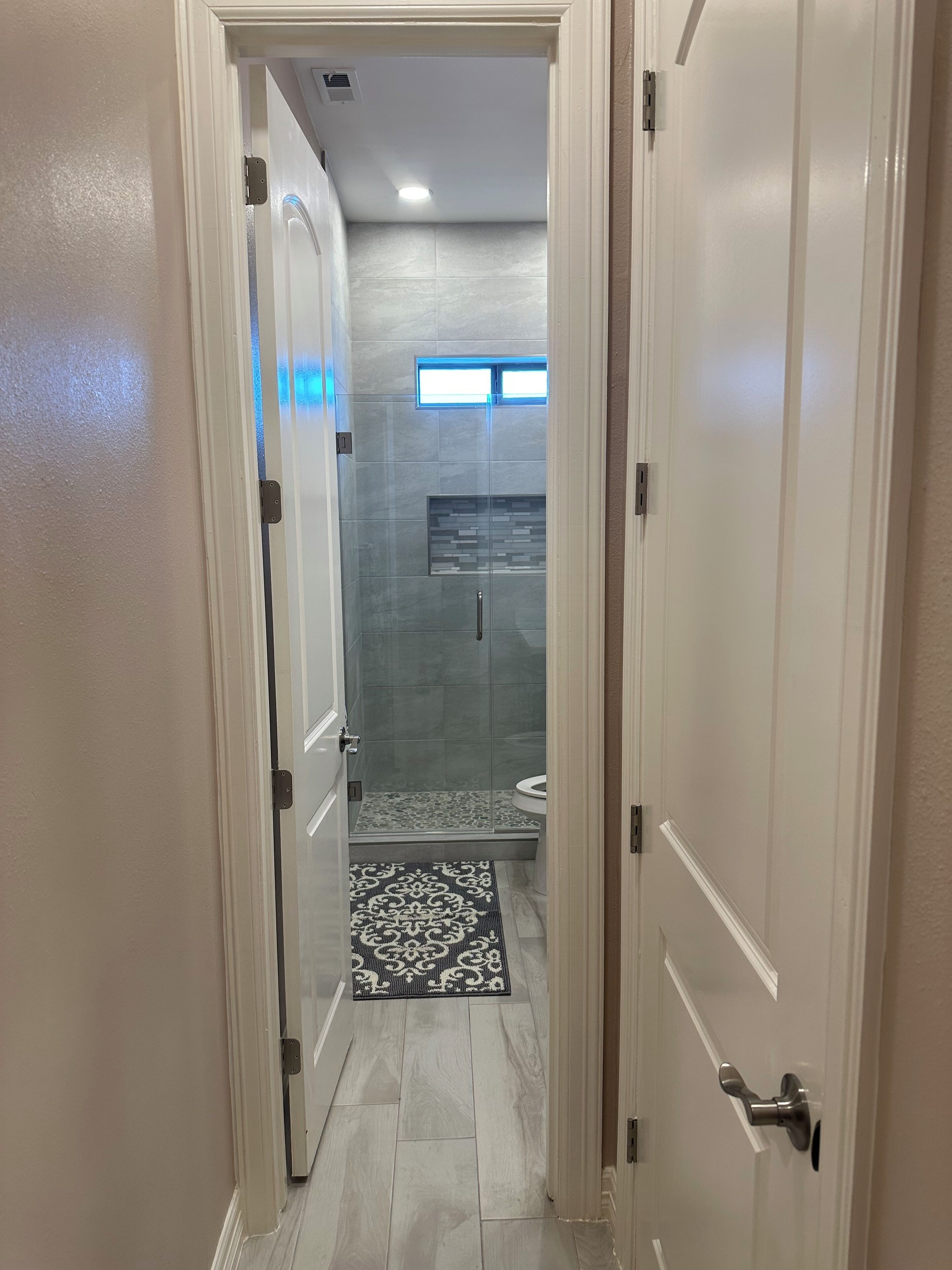 A hallway leading to a bathroom with a glass shower enclosure, a small window, and a patterned rug on the floor.