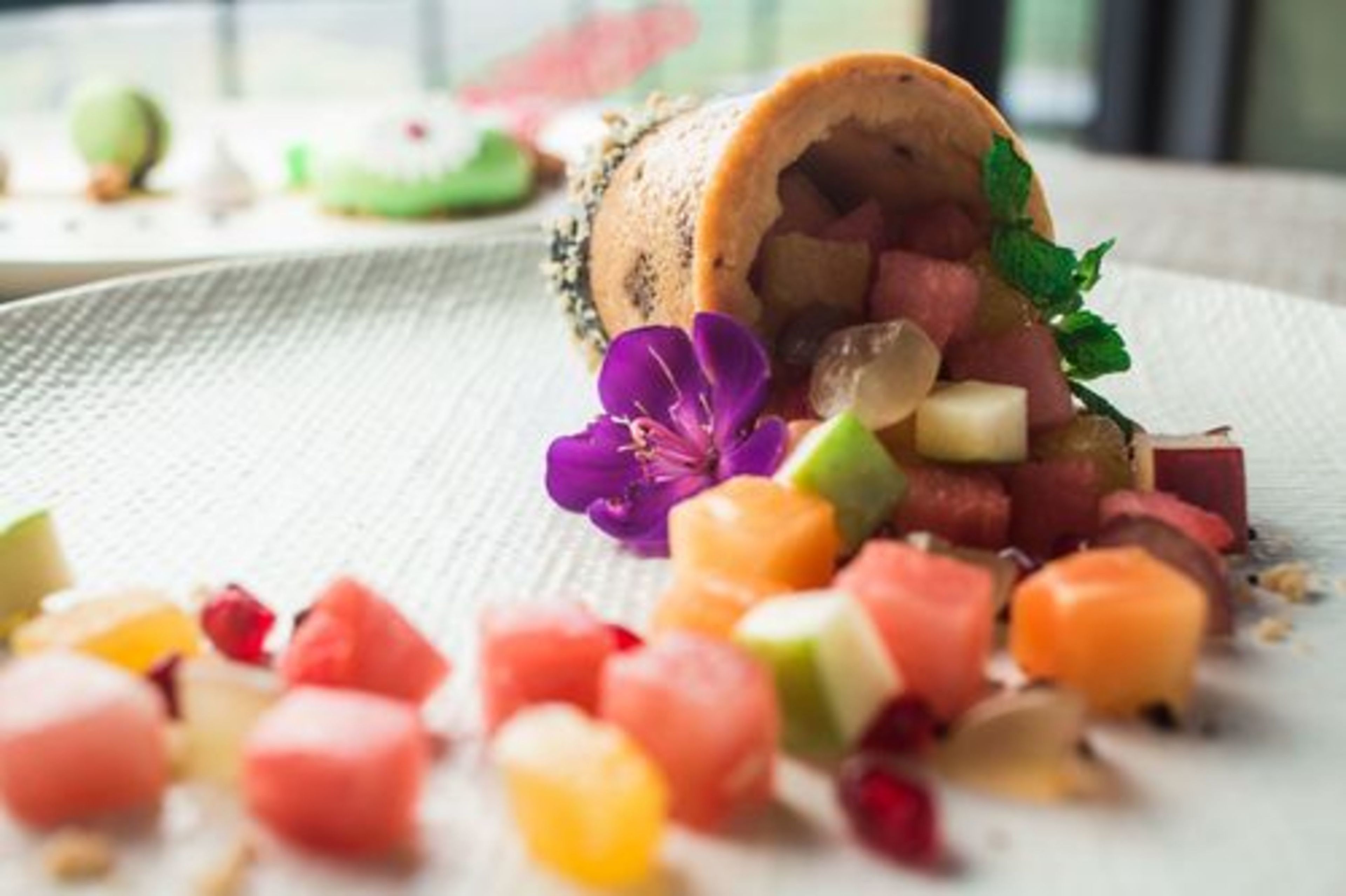 A dessert with a rolled pastry, surrounded by colorful fruit pieces and a purple flower.