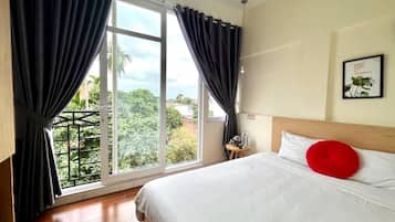 A bedroom with a large window, a bed with white bedding and a red pillow, and a small table with a lamp.