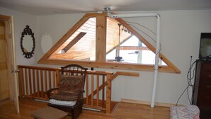 Una habitación con techo inclinado, muebles de madera, una silla con cojín, una mesita y un televisor sobre un mueble de madera.