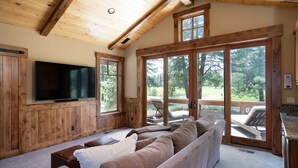 Ein gemütliches Wohnzimmer mit einem großen Flachbildfernseher, Holzwandverkleidung, einer Couch und großen Fenstern mit Blick ins Freie.