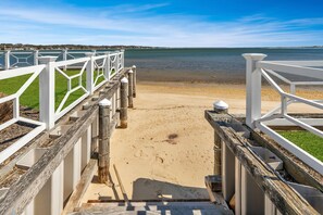 Eine Holzstege führt zu einem Sandstrand mit freiem Blick auf das Wasser und die Küste in der Ferne.