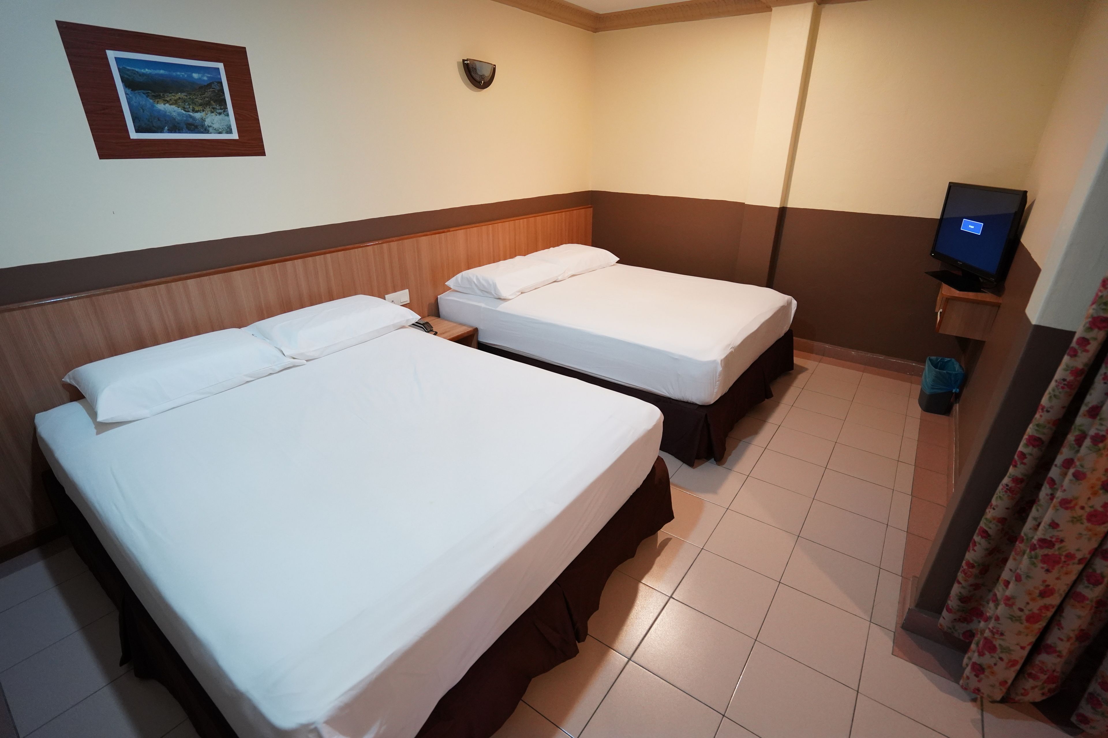 Habitación de hotel con dos camas individuales, televisión, luz empotrada en la pared y una imagen de una playa.